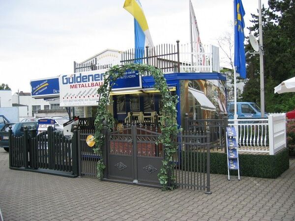 Metallbau-Firma mit dekorativem Tor, Zaun, Firmenlogo und Fahnen auf dem Firmengelände.