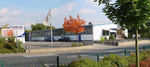 Bürogebäude mit Parkplatz und Firmenlogo Güldener an einer belebten Straße im Gewerbegebiet.