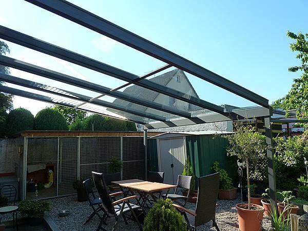 Terrassenüberdachung aus Glas im modernen Garten mit Sitzgruppe und Pflanzen bei Sonnenschein.