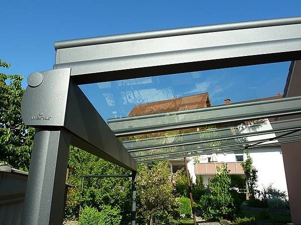 Modernes Terrassendach aus Glas und Aluminium vor Garten und Wohnhaus bei Sonnenschein.