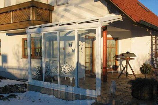 Moderner Wintergarten aus Glas mit weißen Rahmen an Einfamilienhaus im Sonnenlicht.