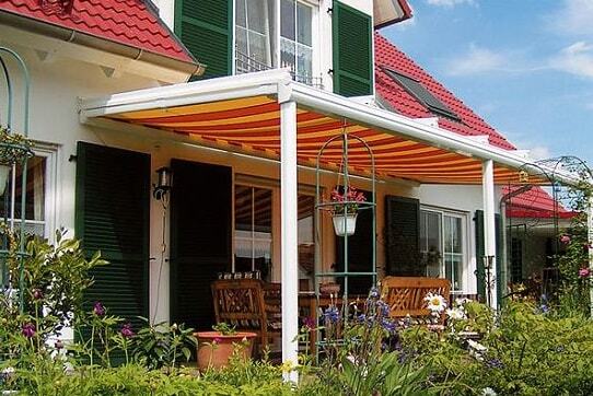 Überdachte Terrasse mit Markise und Gartenmöbeln vor Einfamilienhaus mit Blumenbeet.